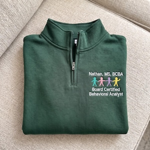 May include: A dark green quarter-zip pullover featuring white embroidered text that reads Nathan, MS, BCBA and Board Certified Behavioral Analyst, accompanied by four small, colorful stick figure icons in teal, pink, purple, and yellow.