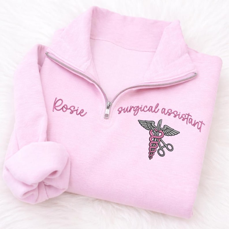 May include: A light pink quarter-zip sweatshirt with the name "Rosie" embroidered on the left chest. The words "surgical assistant" and a medical symbol are embroidered on the right chest. The medical symbol includes a caduceus, wings, and scissors.