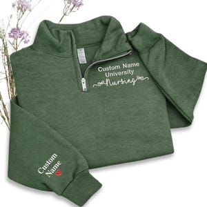 May include: A forest green quarter-zip sweatshirt with white embroidered text that reads "Custom Name University Nursing". The left sleeve has "Custom Name" embroidered with a red heart. The zipper is silver.