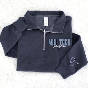 May include: A dark gray quarter-zip sweatshirt with the text "MRI TECH Kayla" in light blue. A stethoscope graphic is on the sleeve. The sweatshirt has a silver zipper and a folded collar.