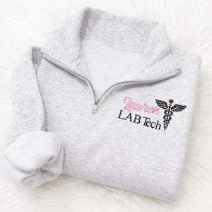 May include: A light gray quarter-zip sweatshirt with a silver zipper. The sweatshirt features the name "Lauren" in pink script, the words "LAB Tech" in black, and a black medical symbol.