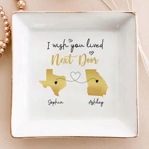May include: White square ceramic trinket dish with gold trim. The dish features gold outlines of the states of Texas and Georgia, connected by a dotted line with hearts. Text reads "I wish you lived Next Door", with names "Sophia" and "Ashley".