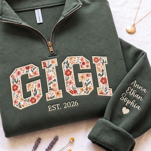 May include: An olive green quarter-zip sweatshirt featuring the word GIGI in large block letters filled with a floral pattern of red, orange, and white flowers. Below the text is EST. 2026. The sleeve is embroidered with the names Anna, Ethan, Sophia.
