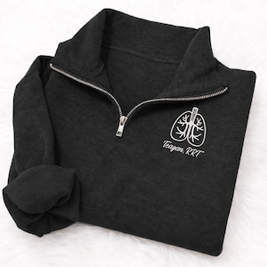May include: Dark gray quarter-zip sweatshirt with a white lung graphic and the text "Teagan, RRT". The sweatshirt has a silver zipper and folded sleeves. The fabric appears soft and comfortable.