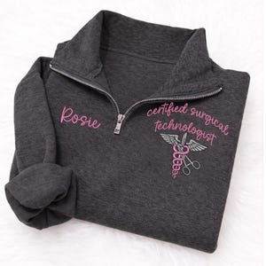 May include: Dark gray quarter-zip sweatshirt with the name "Rosie" embroidered in pink on the left chest. The right chest features the words "certified surgical technologist" and a medical symbol, also in pink.
