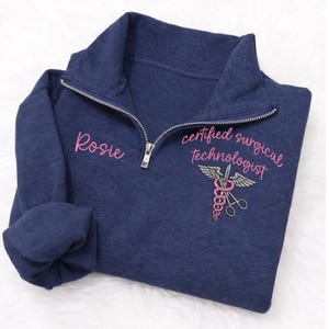 May include: Navy blue quarter-zip sweatshirt with the name "Rosie" embroidered in pink on the left chest. The right chest features a pink and gray medical symbol with the words "certified surgical technologist".