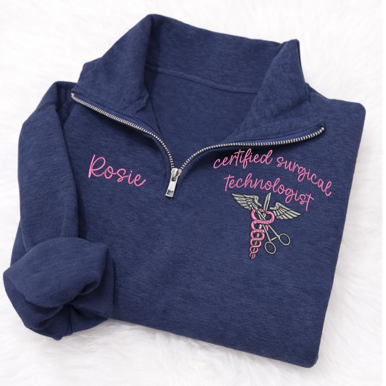 May include: Navy blue quarter-zip sweatshirt with the name "Rosie" embroidered in pink on the left chest. The right chest features a pink and gray medical symbol with the words "certified surgical technologist".