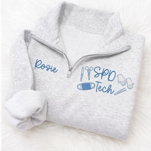 May include: A light gray quarter-zip sweatshirt with the name "Rosie" embroidered in blue on the left chest. The right chest features blue embroidered medical symbols and the text "SPD Tech". The sweatshirt has a silver zipper.