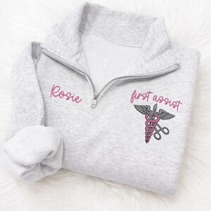 May include: A light gray quarter-zip sweatshirt with the name "Rosie" embroidered in pink on the left chest. The right chest features a pink and gray medical symbol with the words "first assist" in pink.