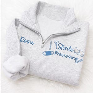May include: A light gray quarter-zip sweatshirt with the name "Rosie" embroidered in blue on the left chest. The right chest features blue embroidered medical-themed graphics and the words "Sterile Processing".