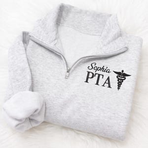May include: A light gray quarter-zip sweatshirt with the name "Sophia" and "PTA" printed in black, along with a caduceus symbol. The sweatshirt has a folded sleeve and a silver zipper.