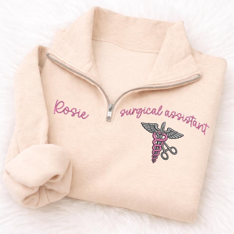 May include: A light beige quarter-zip sweatshirt with the name "Rosie" embroidered in pink script on the left chest. The words "surgical assistant" are embroidered in pink script on the right chest, along with a medical symbol.