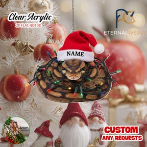 May include: A clear acrylic flat ornament featuring a coiled brown and black patterned snake wearing a red and white Santa hat with the text NAME printed on the brim. The snake is wrapped in a string of colorful Christmas lights.