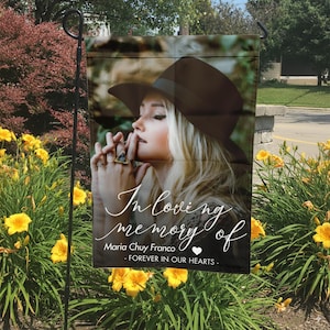 May include: A garden flag featuring a portrait of a woman wearing a hat, with the text "In loving memory of Maria Chuy Franco - Forever in our hearts." The flag is displayed outdoors, surrounded by yellow flowers.