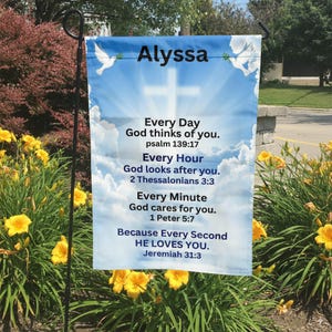 May include: A garden flag with a blue sky background and a white cross. The flag has the name "Alyssa" at the top and inspirational text from the Bible. Two white doves are on the flag. Yellow flowers and green plants are in the foreground.