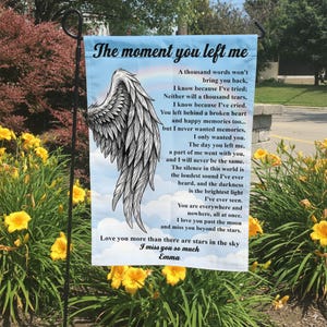 May include: A garden flag with the text "The moment you left me" and a poem about loss. The flag features a large, detailed illustration of a white angel wing against a blue sky background.