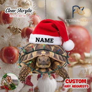 May include: A flat, clear acrylic hanging ornament featuring a tortoise wearing a red and white Santa hat with the text NAME printed on the brim. The tortoise is wrapped in a string of colorful Christmas lights. A small inset shows a 4mm thickness.