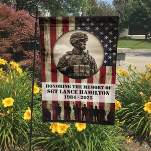 May include: A garden flag featuring an American flag design with a sepia-toned portrait of a soldier in a circular frame. The flag reads "Honoring the Memory of Sgt. Lance Hamilton 1984-2025" with a silhouette of soldiers at the bottom.