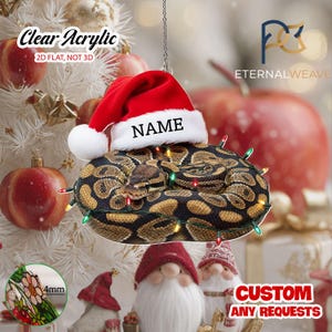 May include: A clear acrylic 2D flat hanging ornament featuring a coiled ball python wearing a red and white Santa hat with the text NAME printed on the brim. The snake is wrapped in a string of small multicolored Christmas lights.