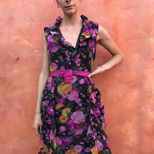 May include: A sleeveless maxi dress with a floral print in shades of pink, purple, orange, and yellow on a black background. The dress has a ruffled neckline and a pink belt.