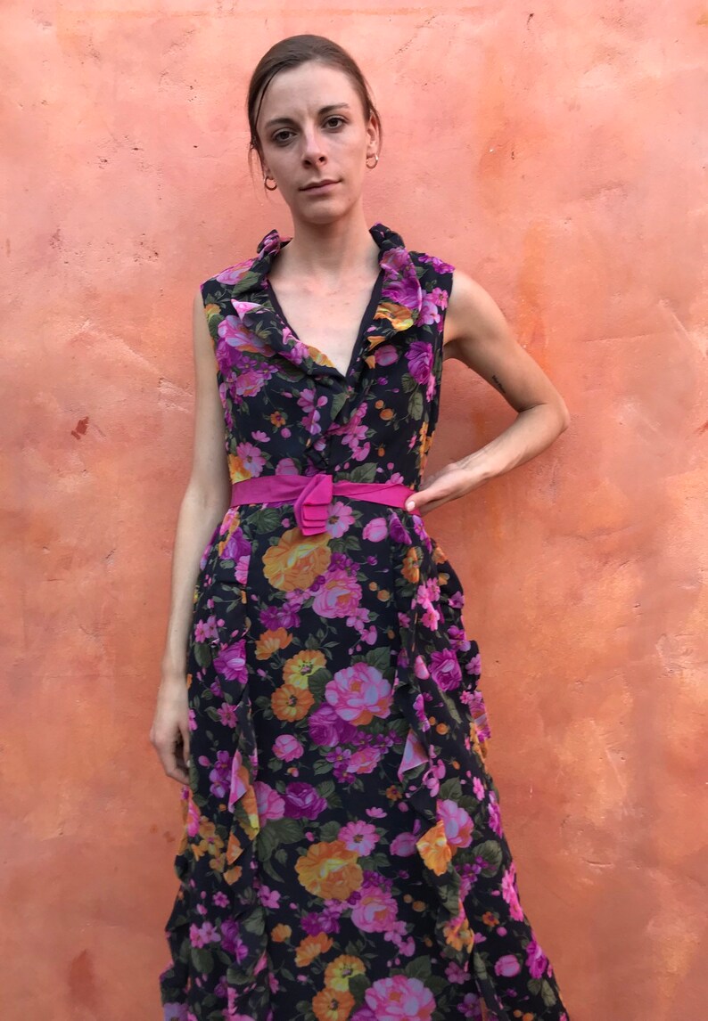 May include: A sleeveless maxi dress with a floral print in shades of pink, purple, orange, and yellow on a black background. The dress has a ruffled neckline and a pink belt.