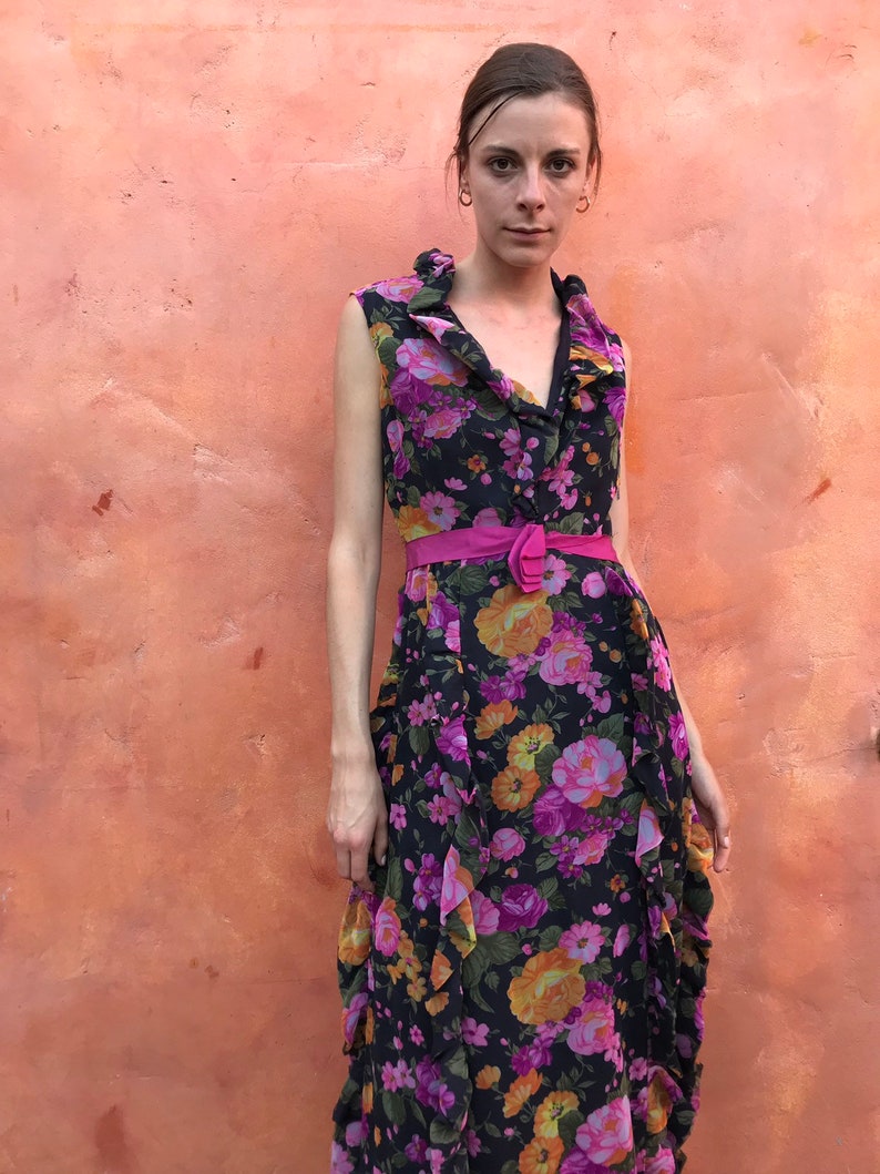 May include: A sleeveless maxi dress with a floral print in shades of pink, orange, and yellow on a black background. The dress has a ruffled neckline and a pink belt.