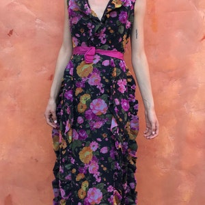 May include: A woman wearing a long, black floral dress with a pink belt. The dress has a ruffled neckline and a flowing skirt. She is wearing black sandals.