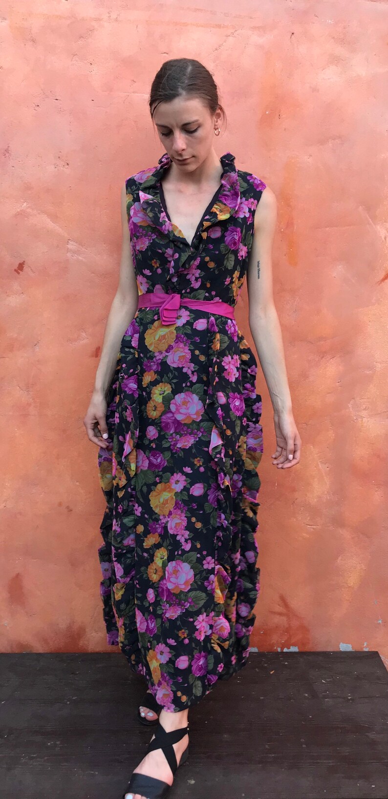 May include: A woman wearing a long, black floral dress with a pink belt. The dress has a ruffled neckline and a flowing skirt. She is wearing black sandals.
