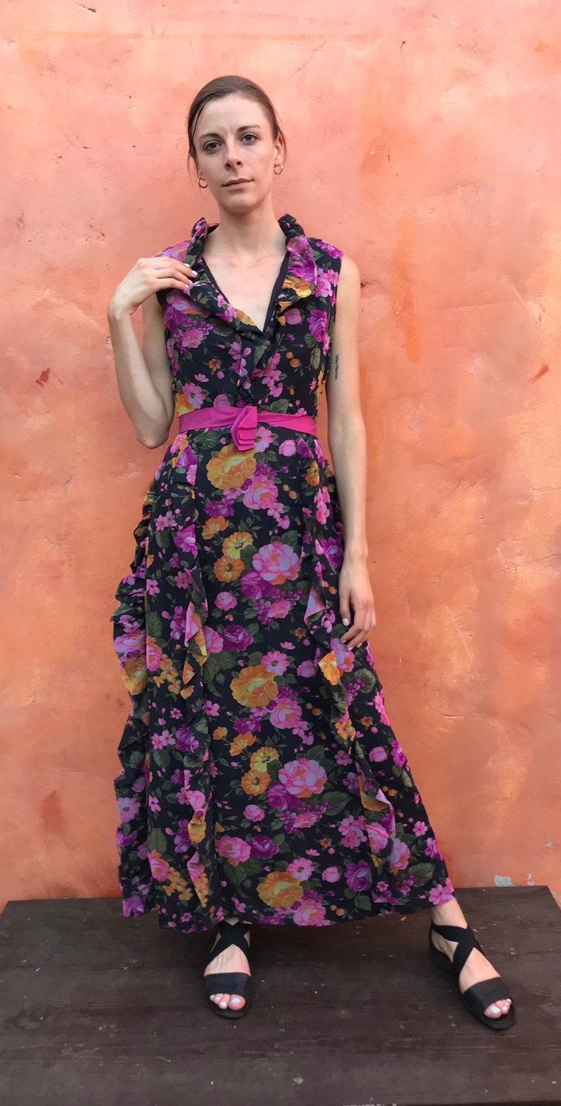 May include: A woman wearing a long, black floral dress with a pink ruffled collar and a pink belt. She is wearing black sandals with straps.