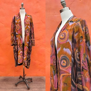 May include: A long, open-front kimono-style jacket with a vibrant abstract pattern in shades of orange, pink, and brown. The jacket is displayed on a mannequin against an orange background. The design features circular and geometric shapes.