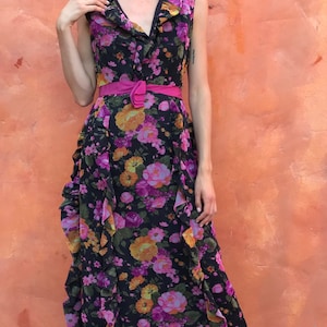 May include: A woman wearing a black maxi dress with a pink floral print and a pink belt. She is standing in front of a peach-coloured wall.