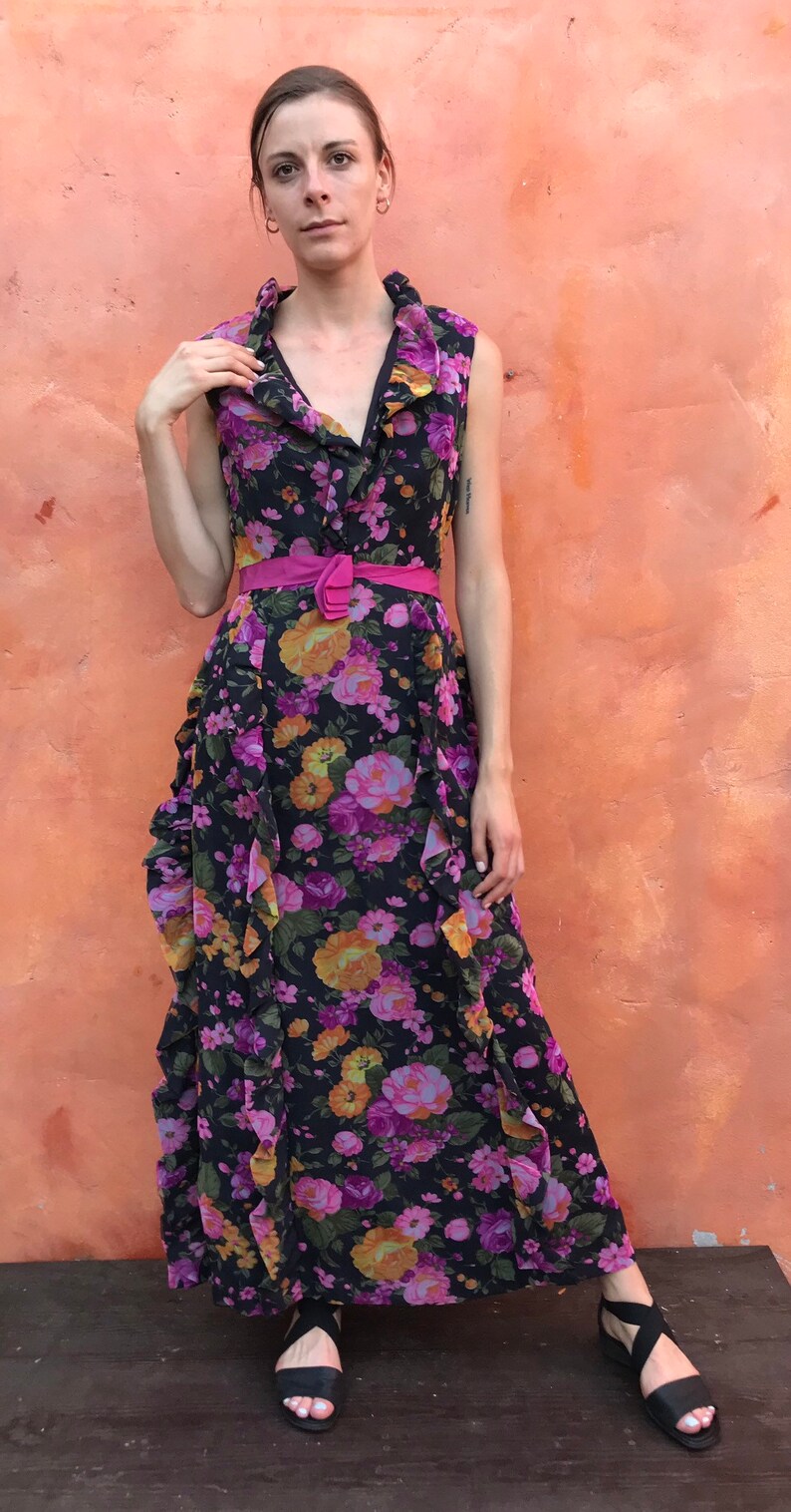 May include: A woman wearing a black maxi dress with a pink floral print and a pink belt. She is standing in front of a peach-coloured wall.