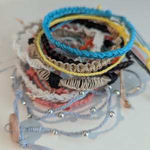 May include: A collection of handmade bracelets in various colors, including blue, yellow, black, and beige. Some bracelets feature braided designs, while others have small silver beads and a button closure. The bracelets are stacked together, showcasing a variety of styles.