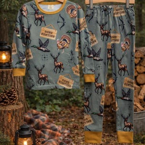 Puede incluir: Un conjunto de pijama de dos piezas con temática de caza. El conjunto incluye una camiseta de manga larga y pantalones, ambos en color verde apagado con un patrón repetido de ciervos, patos, pavos, arcos y letreros que dicen "Gone Hunting". Los puños y el cuello están ribeteados en amarillo.
