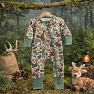 Könnte beinhalten: Ein Baby-Schlafanzug mit Camouflage-Muster und Reißverschluss, mit braunen, grünen und beigen Formen sowie Hirschsilhouetten. Der Schlafanzug hat grüne Bündchen und Verzierungen. Er hängt an einem Holzbügel vor einem Waldhintergrund.