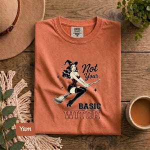 May include: A burnt orange crew neck t-shirt featuring a black graphic of a pin-up style witch riding a broomstick. The text reads Not Your BASIC WITCH. The shirt has a Comfort Colors brand label at the inner neckline and is size M.