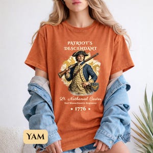 Puede incluir: Camiseta naranja con una ilustraci&oacute;n hist&oacute;rica de un hombre con uniforme colonial sosteniendo un rifle. El texto "PATRIOT'S DESCENDANT" est&aacute; encima de la imagen, con "Lt. Nathaniel Carter" y "1776" debajo.