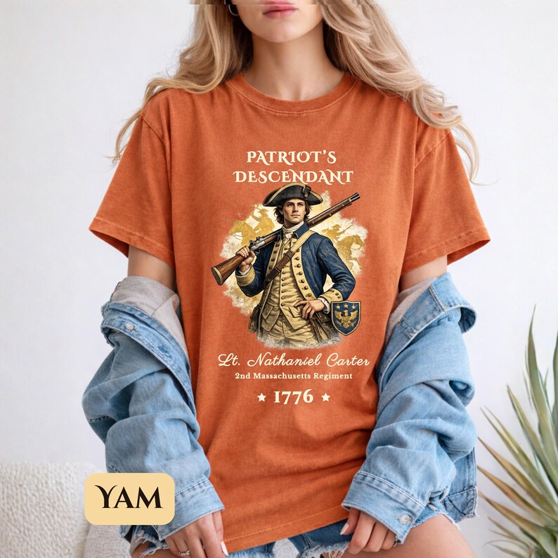 Puede incluir: Camiseta naranja con una ilustraci&oacute;n hist&oacute;rica de un hombre con uniforme colonial sosteniendo un rifle. El texto "PATRIOT'S DESCENDANT" est&aacute; encima de la imagen, con "Lt. Nathaniel Carter" y "1776" debajo.