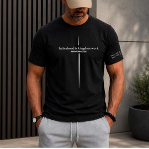 May include: Black t-shirt with the text "fatherhood is kingdom work" and "PROVERBS 22:6" in white. A vertical white line design is centered on the shirt. The right sleeve has the text "Olivia EST. 20" and "Jasper EST. 20".
