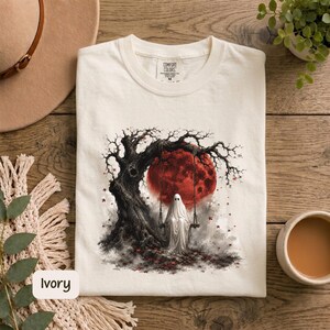 May include: An ivory t-shirt featuring a graphic print of a white ghost sitting on a swing attached to a dark, gnarled tree. Behind the tree is a large, textured red moon. The shirt has a crew neckline and a Comfort Colors brand label at the neck.