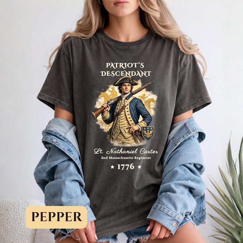 Puede incluir: Camiseta gris oscuro con el texto "PATRIOT'S DESCENDANT" y una ilustraci&oacute;n de un hombre con abrigo azul sosteniendo un rifle. El texto "Lt. Nathaniel Carter 2nd Massachusetts Regiment" y el a&ntilde;o "1776" tambi&eacute;n est&aacute;n impresos.