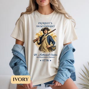 Puede incluir: Camiseta de color marfil con un gr&aacute;fico de un soldado colonial sosteniendo un rifle, con el texto "PATRIOT'S DESCENDANT" arriba y "Lt. Nathaniel Carter" abajo. La camiseta tambi&eacute;n incluye el texto "2nd Massachusetts Regiment" y "1776".