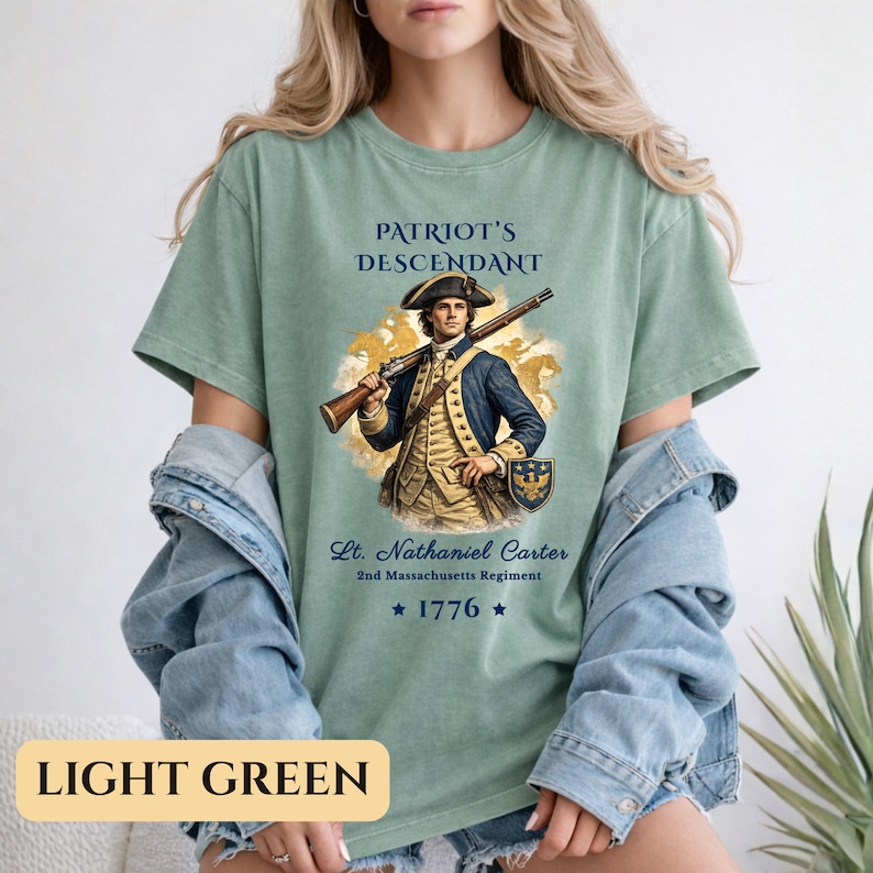 Puede incluir: Camiseta verde claro con el texto "PATRIOT'S DESCENDANT" sobre una ilustraci&oacute;n de un hombre con uniforme colonial sosteniendo un rifle. Debajo est&aacute; el texto "Lt. Nathaniel Carter 2nd Massachusetts Regiment ★ 1776 ★". La parte inferior de la camiseta tiene el texto "LIGHT GREEN".