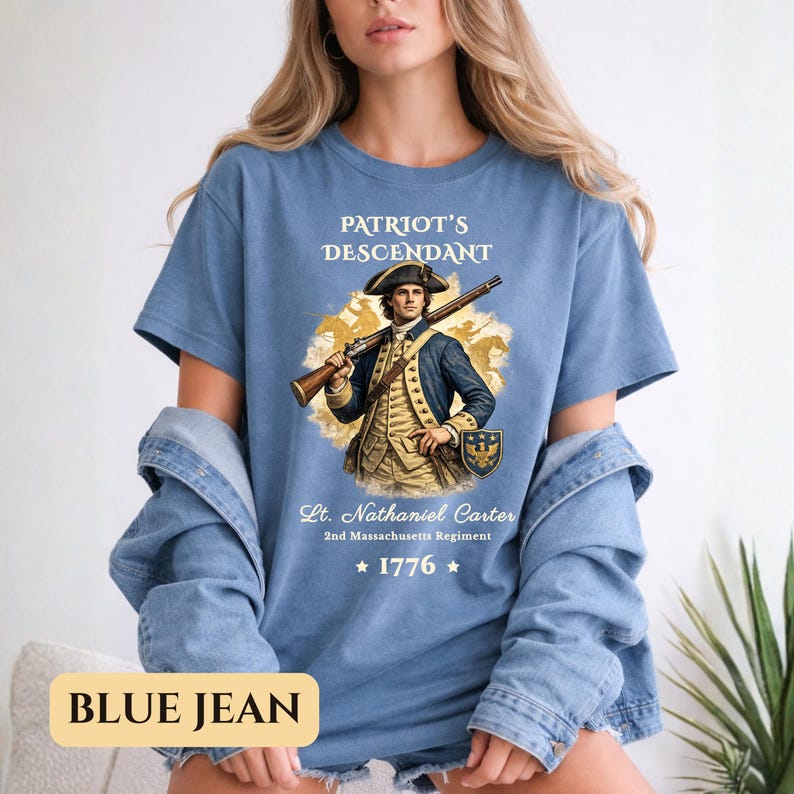 Puede incluir: Camiseta azul con el texto "PATRIOT'S DESCENDANT" y una ilustraci&oacute;n de un hombre con uniforme colonial sosteniendo un rifle. El texto "Lt. Nathaniel Carter 2nd Massachusetts Regiment" y el a&ntilde;o "1776" tambi&eacute;n est&aacute;n impresos.