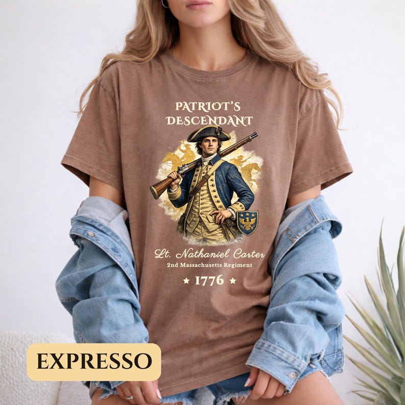 Puede incluir: Camiseta marr&oacute;n con el texto "PATRIOT'S DESCENDANT" y una ilustraci&oacute;n de un soldado con un rifle. El texto "Lt. Nathaniel Carter 2nd Massachusetts Regiment" y el a&ntilde;o "1776" tambi&eacute;n est&aacute;n impresos.