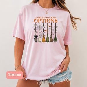 May include: A light pink t-shirt featuring a graphic of six different broomsticks arranged in a row. Above the brooms, the text reads A GIRL'S GOTTA HAVE OPTIONS in orange. The design includes spiderwebs and small orange bats in the background.