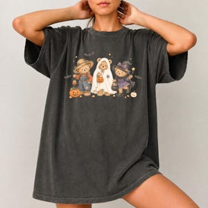 May include: A charcoal gray short-sleeve t-shirt featuring a graphic print of three teddy bears dressed for Halloween. One bear wears a straw hat and overalls, one is dressed as a ghost, and one wears a witch costume with a purple hat and wand.