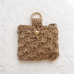 May include: A square-shaped pouch crafted from metallic gold yarn in a textured crochet stitch. The pouch features a gold-toned metal ring attached to the top center and a single gold-colored snap button closure centered on the front flap.