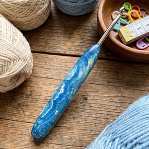 May include: A blue and white marbled crochet hook with a silver metal hook. The hook is next to balls of cream and light blue yarn, a wooden bowl with stitch markers, and a measuring tape. The items are on a wooden surface.