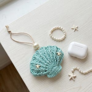 May include: A crocheted teal seashell-shaped case with attached pearl and string, adorned with small starfish. A white wireless earbud case, a pearl bracelet, and a pearl necklace with starfish accents are also present.